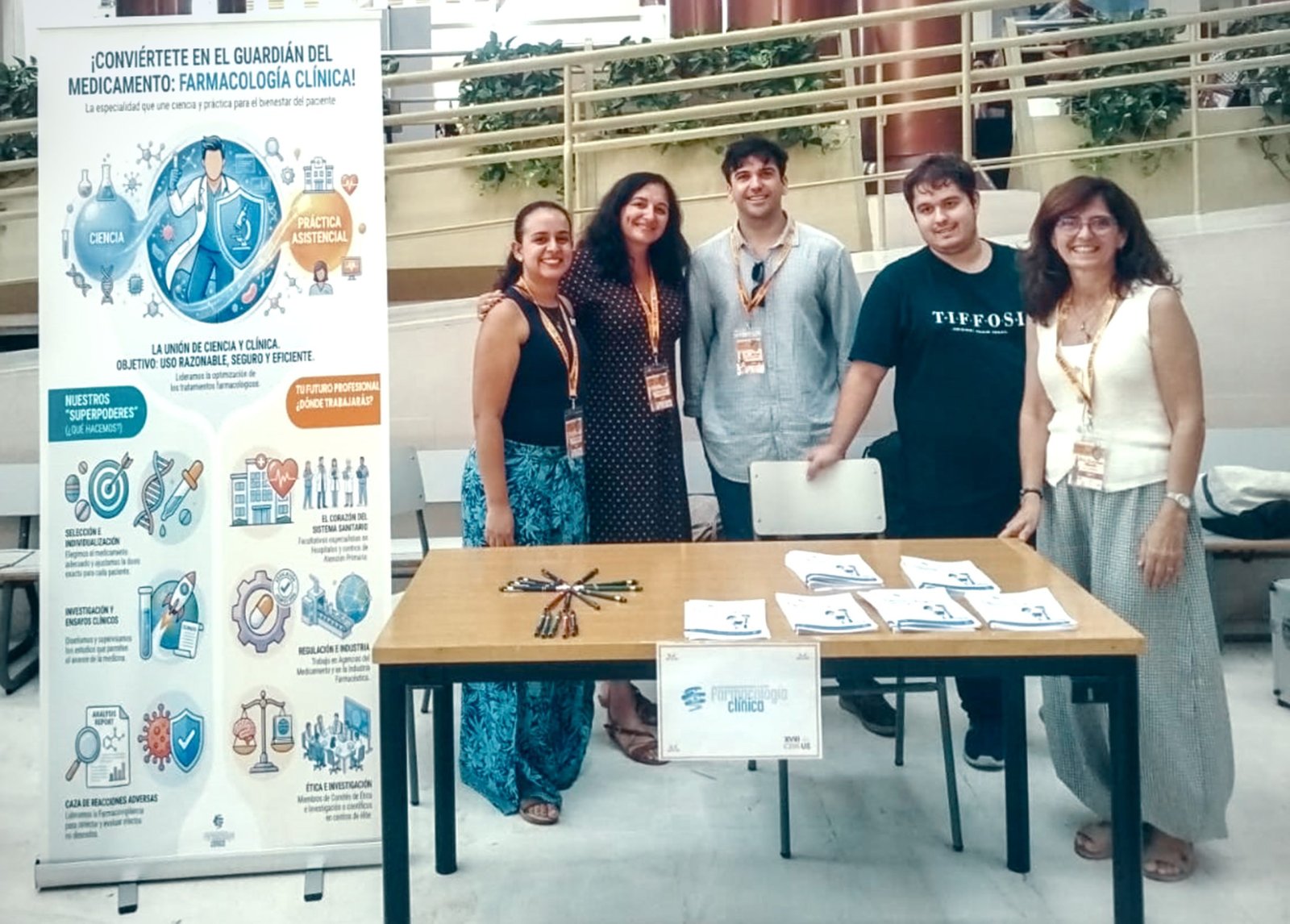 Cinco personas sonríen tras una mesa con folletos y bolígrafos, junto a un cartel sobre Farmacología Clínica.