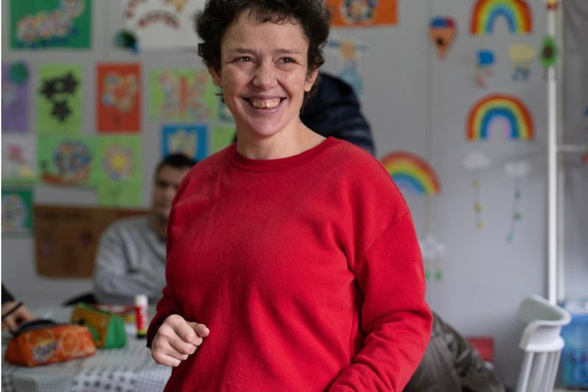Una mujer sonriente con jersey rojo posa en una sala con dibujos infantiles de arcoíris en la pared y otras personas al fondo.