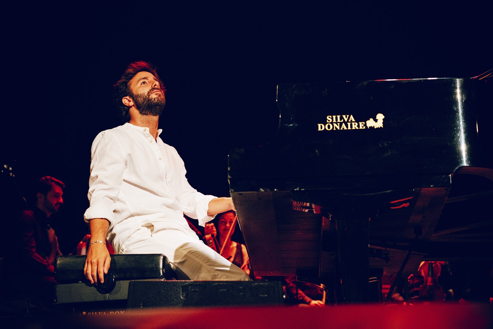 Un hombre con barba y camisa blanca sentado al piano de cola en un escenario oscuro, con músicos al fondo.