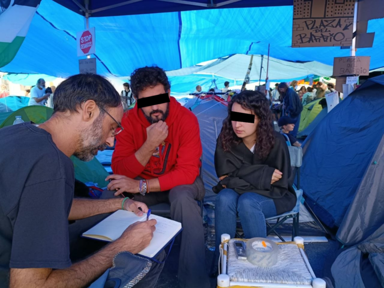 Un hombre con libreta y bolígrafo toma notas sentado frente a dos personas con los ojos tapados, bajo una carpa azul junto a tiendas de campaña.
