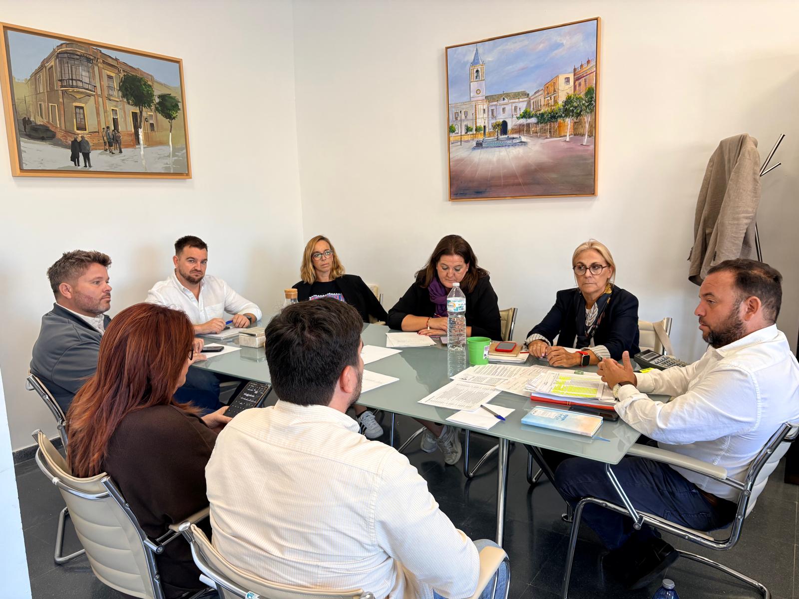 Siete personas sentadas alrededor de una mesa de reuniones con documentos, en una sala con dos cuadros de edificios en la pared.