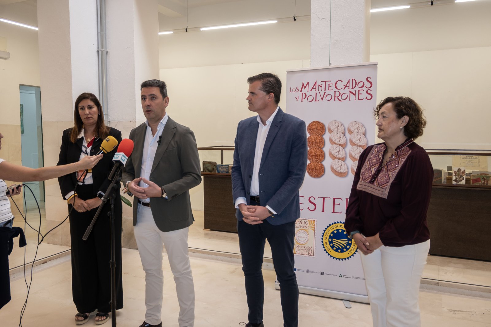 Tres personas de pie junto a un cartel de mantecados y polvorones mientras un micrófono les entrevista en una sala.
