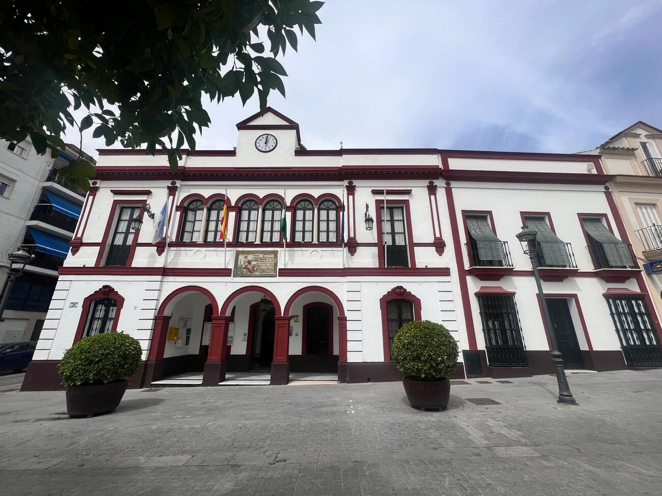 Edificio de dos plantas blanco con remates granates, arcos en la planta baja, ventanas enrejadas, reloj y banderas en la fachada, ante una plaza empedrada.
