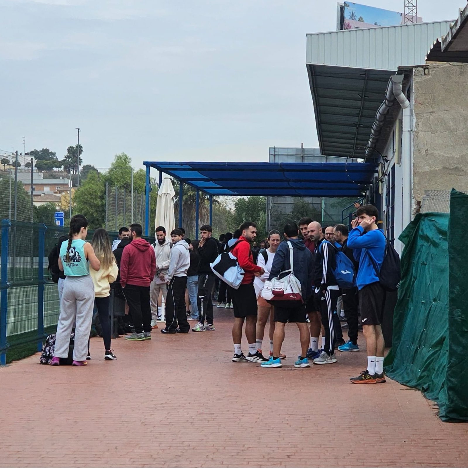 Un grupo de personas con ropa deportiva espera de pie junto a una pista deportiva con valla metálica y una marquesina azul.