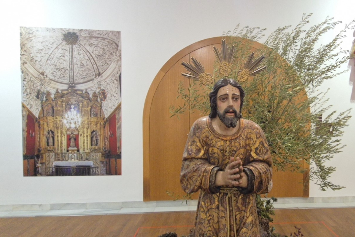 Una talla de Cristo con las manos juntas y ropaje dorado, ante un panel de madera y una foto de un retablo.