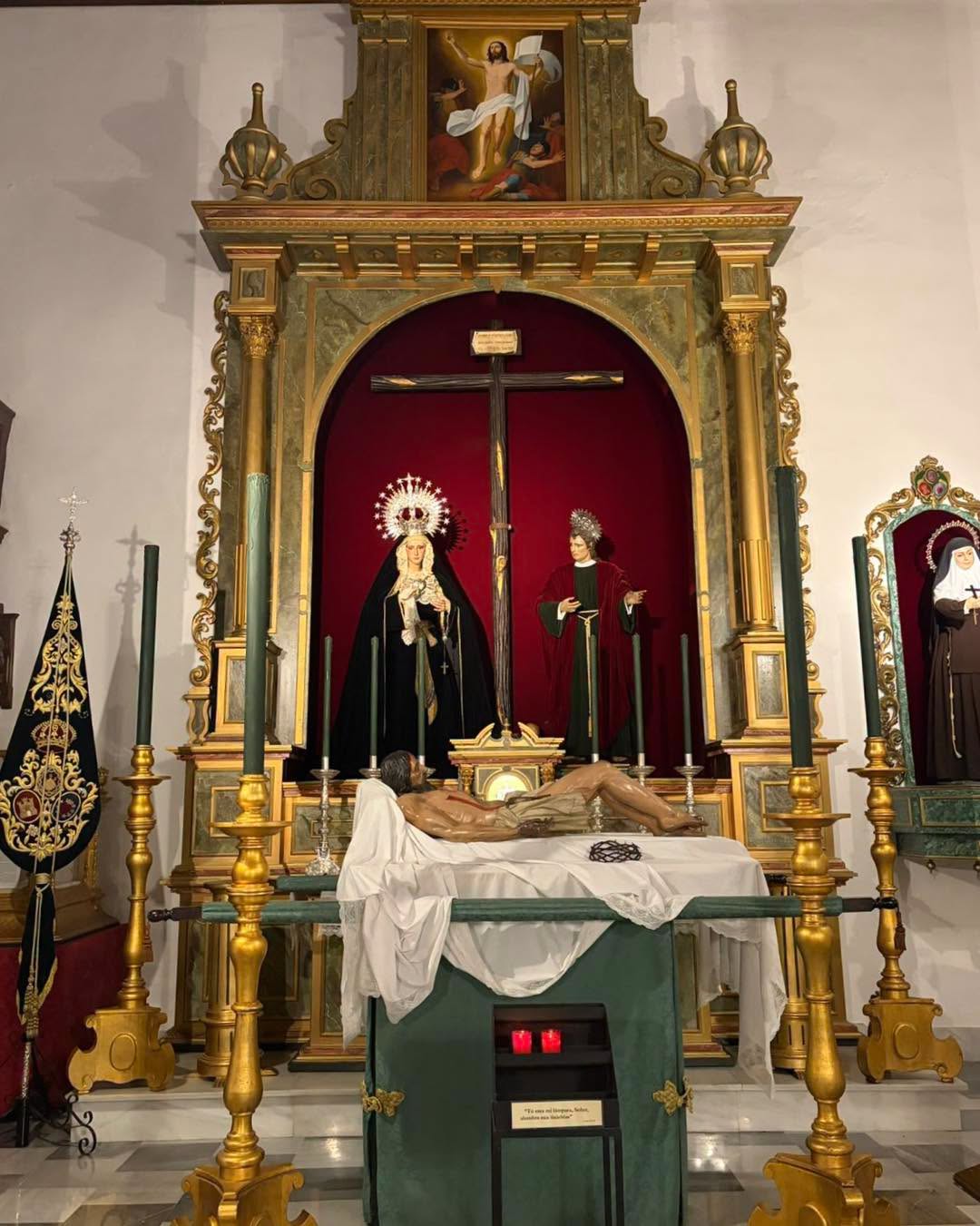 Un retablo dorado con un crucificado, una Virgen dolorosa y un santo, y ante él una imagen yacente sobre un túmulo verde.