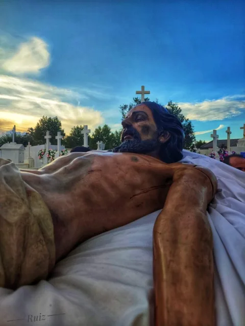 Una talla de Cristo yacente sobre sábanas blancas, con cruces y tumbas de un cementerio al fondo bajo un cielo azul.