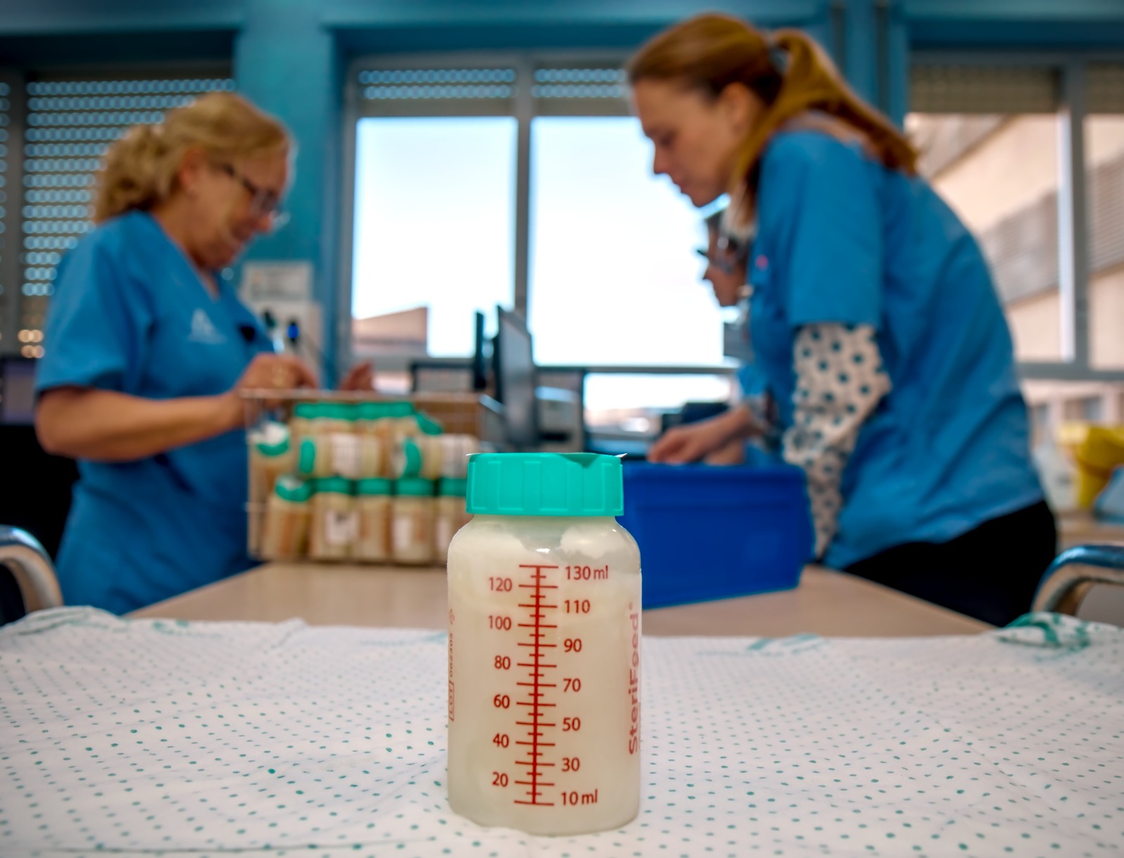 Un biberón graduado con leche en primer plano sobre una mesa y, al fondo desenfocadas, dos personas con pijama azul junto a varios botes.