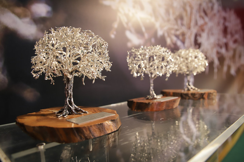 Tres esculturas de árboles de metal brillante sobre bases de madera, alineadas sobre una superficie de cristal.