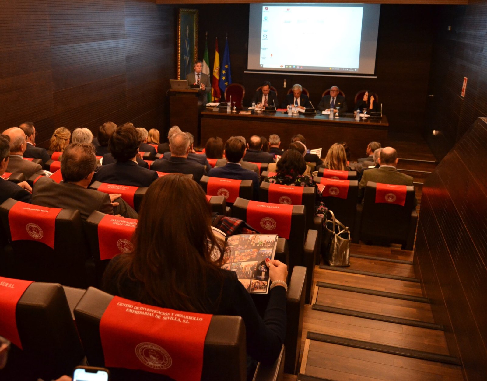Público sentado en un salón de actos con ponentes en una mesa y una pantalla de proyección al fondo.