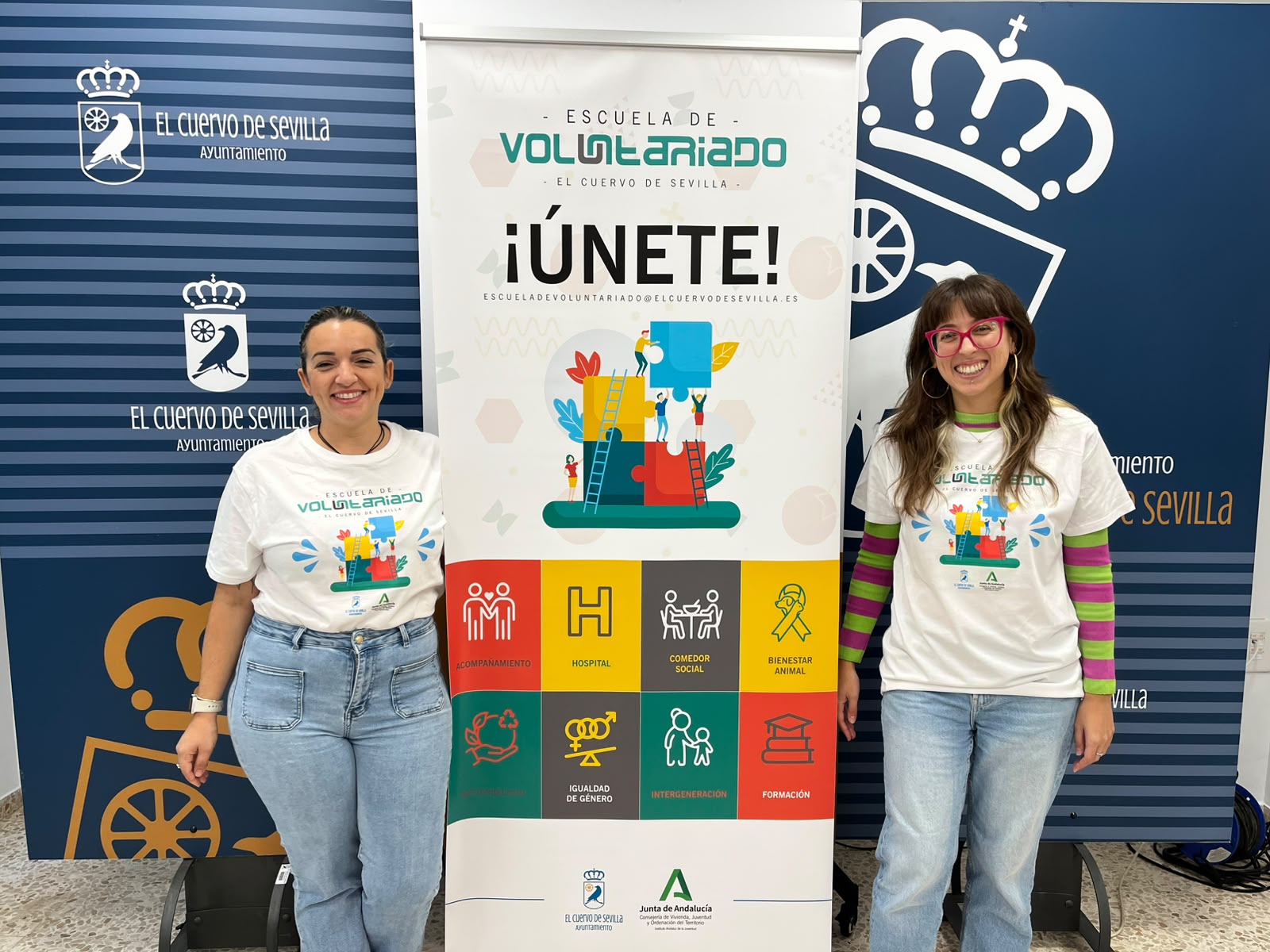 Dos mujeres sonrientes con camisetas blancas posan junto a un cartel de una escuela de voluntariado.