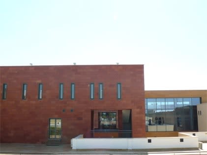 Edificio moderno de ladrillo rojizo con ventanas verticales estrechas y una zona acristalada, bajo un cielo despejado.
