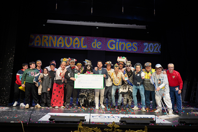 Un grupo de personas disfrazadas posa en un escenario con un cartel de Carnaval de Gines 2026 y un cheque grande.