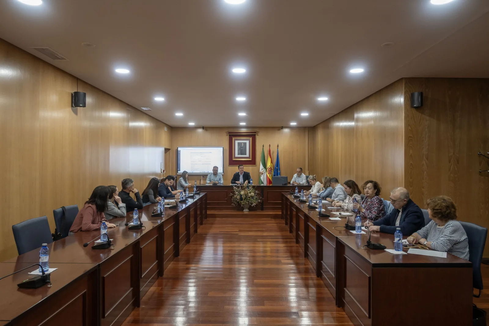 Varias personas sentadas alrededor de una gran mesa de reuniones de madera en una sala con paredes de madera, pantalla y banderas al fondo.